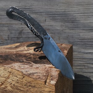 K&ouml;nnte beinhalten: Ein handgeschmiedetes Messer mit einem gebogenen Griff und einer scharfen Klinge. Das Messer liegt auf einem St&uuml;ck Holz.