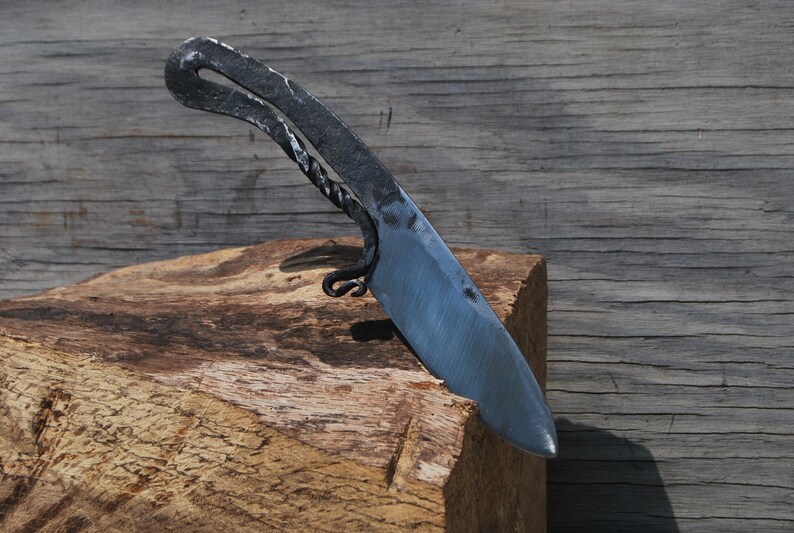 K&ouml;nnte beinhalten: Ein handgeschmiedetes Messer mit einem gebogenen Griff und einer scharfen Klinge. Das Messer liegt auf einem St&uuml;ck Holz.
