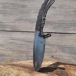 K&ouml;nnte beinhalten: Ein handgeschmiedetes Messer mit einem gedrehten Griff und einer scharfen Klinge. Das Messer liegt auf einem St&uuml;ck Holz.