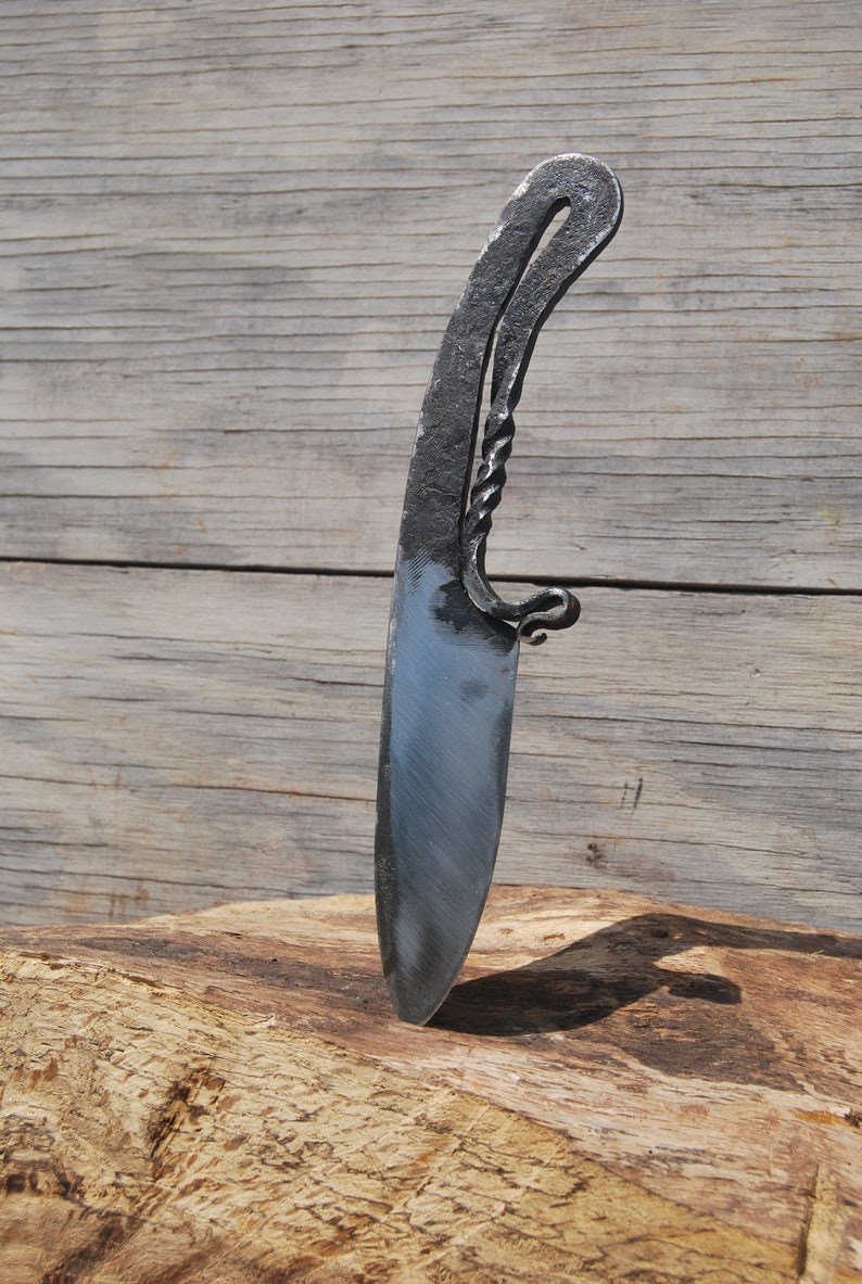 K&ouml;nnte beinhalten: Ein handgeschmiedetes Messer mit einem gedrehten Griff und einer scharfen Klinge. Das Messer liegt auf einem St&uuml;ck Holz.