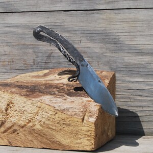 K&ouml;nnte beinhalten: Ein handgeschmiedetes Messer mit einem gebogenen Griff und einer scharfen Klinge, das auf einem St&uuml;ck grob behautem Holz liegt.