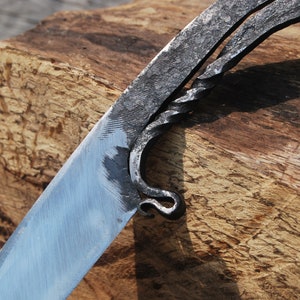 K&ouml;nnte beinhalten: Nahaufnahme eines handgefertigten Messers mit einem gedrehten Metallgriff. Die Klinge ist poliert und der Griff ist rau und strukturiert. Das Messer liegt auf einem St&uuml;ck Holz.