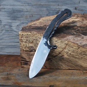 K&ouml;nnte beinhalten: Ein handgeschmiedetes Messer mit einem einzigartigen, gebogenen Griff und einer scharfen, polierten Klinge. Das Messer liegt auf einem St&uuml;ck grob behautem Holz.