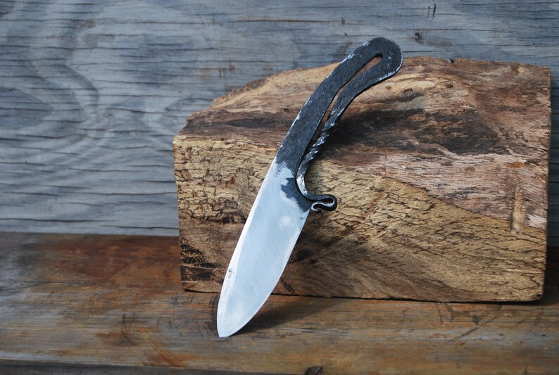 K&ouml;nnte beinhalten: Ein handgeschmiedetes Messer mit einem einzigartigen, gebogenen Griff und einer scharfen, polierten Klinge. Das Messer liegt auf einem St&uuml;ck grob behautem Holz.
