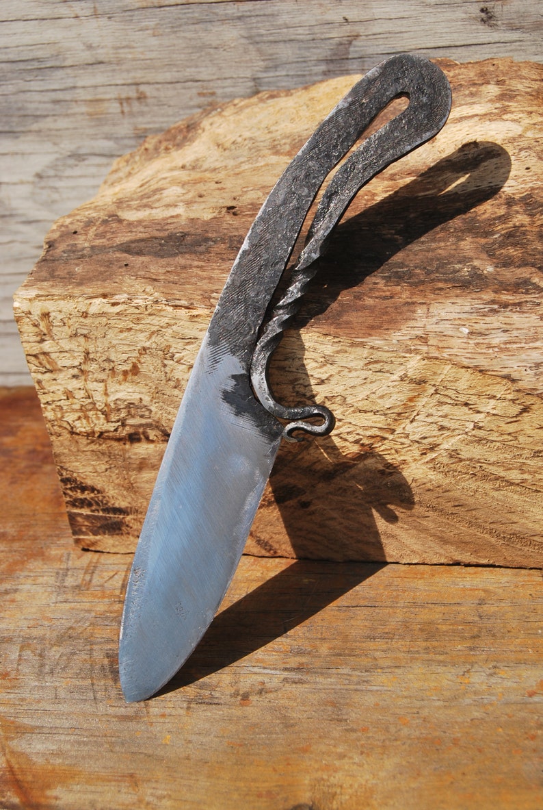K&ouml;nnte beinhalten: Ein handgeschmiedetes Messer mit einem gedrehten Griff und einer scharfen Klinge. Das Messer liegt auf einem St&uuml;ck Holz.