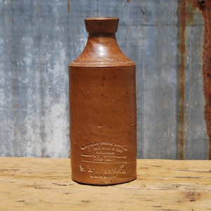 May include: A brown stoneware bottle with a cylindrical body and a narrow neck. The bottle has a textured surface and embossed text. The background is a weathered metal sheet and a wooden surface.