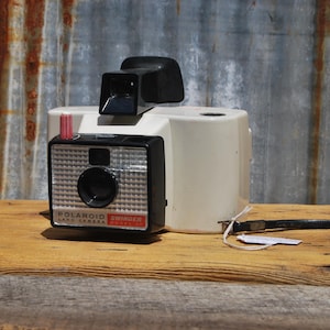May include: A white and black Polaroid Swinger Land Camera with a red and white flash cube. The camera is sitting on a wooden surface.
