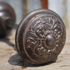 May include: A dark brown, weathered metal door knob featuring a circular shape with an embossed floral and leaf pattern surrounding a central raised dot. The knob is attached to a metal spindle and a matching circular backplate, showing signs of age.