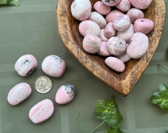 May include: A heart-shaped wooden bowl filled with pink and white tumbled stones. Several stones are scattered around the bowl. A quarter is next to the stones. Green ivy leaves are in the background.