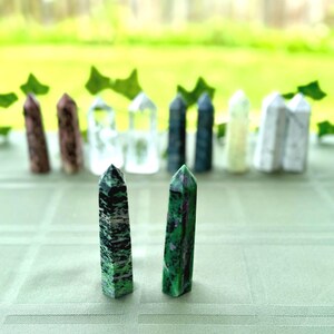 May include: Two green and black gemstone obelisks are in the foreground, with a variety of other gemstone obelisks in the background. The obelisks are on a light green surface, with a blurred green background.