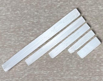 May include: Six white selenite crystal wands of varying lengths are arranged diagonally on a neutral-toned surface. The wands have a translucent, crystalline appearance, with visible striations along their length. The crystals are a natural, earthy product.