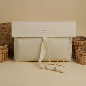 Op de afbeelding: Een crèmekleurige canvas tas met een klepsluiting en een lange, crèmekleurige lintsluiting. Op de tas staat het woord "Hastmala" gedrukt op een klein label. Gevlochten manden staan op de achtergrond.