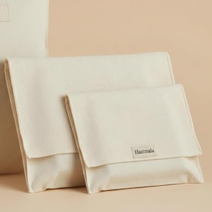 Pode incluir: Bolsas de lona bege claro em vários tamanhos. As bolsas têm formato retangular com fecho de aba. O nome da marca "Hastmala" está impresso em uma pequena etiqueta na frente da menor bolsa. O fundo é de cor bege claro sólido.
