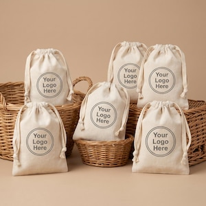 Könnte beinhalten: Eine Anordnung von beigefarbenen Kordelzugbeuteln mit dem Aufdruck "Your Logo Here" in und um geflochtene Körbe. Die Beutel bestehen aus einem Naturstoff und eignen sich für Branding- oder Verpackungszwecke.