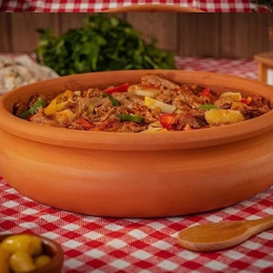 Puede incluir: Una olla de barro llena de un guiso de carne y verduras, incluyendo pimientos rojos y hierbas verdes. La olla está sobre un mantel a cuadros rojos y blancos, con una cuchara de madera cerca. Un tema culinario.