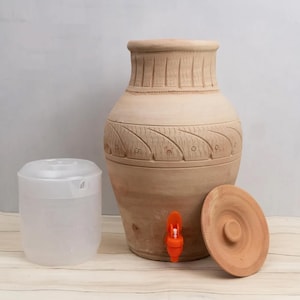 Może przedstawiać: Duży, nieszkliwiony dozownik wody z terakoty z pomarańczowym kranem i pasującą pokrywą. Po lewej stronie znajduje się przezroczysty plastikowy pojemnik z białą pokrywką. Dozownik ma rzeźbione wzory liści i linii.