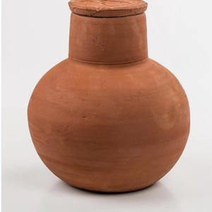 May include: A terracotta-coloured, round-bodied jar with a cylindrical neck and a flat, circular lid. The jar has a smooth, unglazed surface and a rustic, handmade appearance. The lid sits flush on the neck, creating a sealed container.