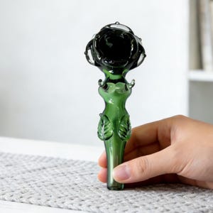 Puede incluir: Una pipa de vidrio verde con un cuenco oscuro en forma de flor. El tallo es de un verde más claro con detalles en forma de hojas. La pipa se sostiene en una mano, mostrando su diseño y artesanía. El fondo es blanco neutro.