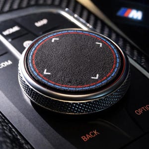 May include: Close-up of a car's control knob. The black, textured knob has a silver rim with red and blue accents. White directional arrows are on the knob's surface. The control panel is black with the letters "HOME", "BACK", and "OPTION".