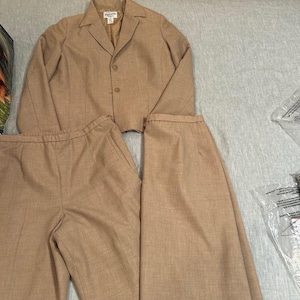 Op de afbeelding: Een beige tweedelig pak, bestaande uit een blazer en een broek. De blazer heeft een reverskraag en drie knopen. De broek heeft rechte pijpen. Het pak is gemaakt van een textuurstof en ligt op een lichtgrijze ondergrond.