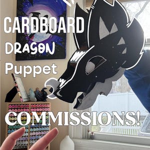 May include: A black and white cardboard dragon puppet with the text "CARDBOARD DRAGON PUPPET COMMISSIONS!" in white. The puppet is held up in front of a window. A painting of a night sky with a moon is in the background.