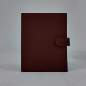 Peut inclure: Un journal en cuir marron au design simple. La couverture est d'une couleur marron riche et unie avec une fermeture à languette assortie sur le côté droit. Le journal est rectangulaire et se tient debout sur une surface blanche.