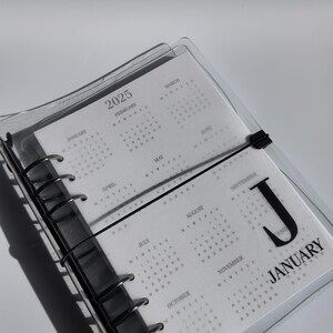 May include: A clear plastic-covered planner for 2025, with a black metal binder. The planner displays monthly calendars. The word "January" is visible on one page. A black elastic band secures the planner.