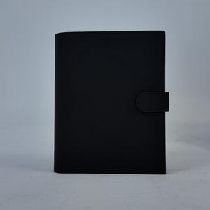 Peut inclure: Un journal noir rectangulaire avec une couverture textur&eacute;e et une fermeture magn&eacute;tique. Le journal est pos&eacute; verticalement sur un fond uni gris clair. La couverture a une finition lisse et mate.