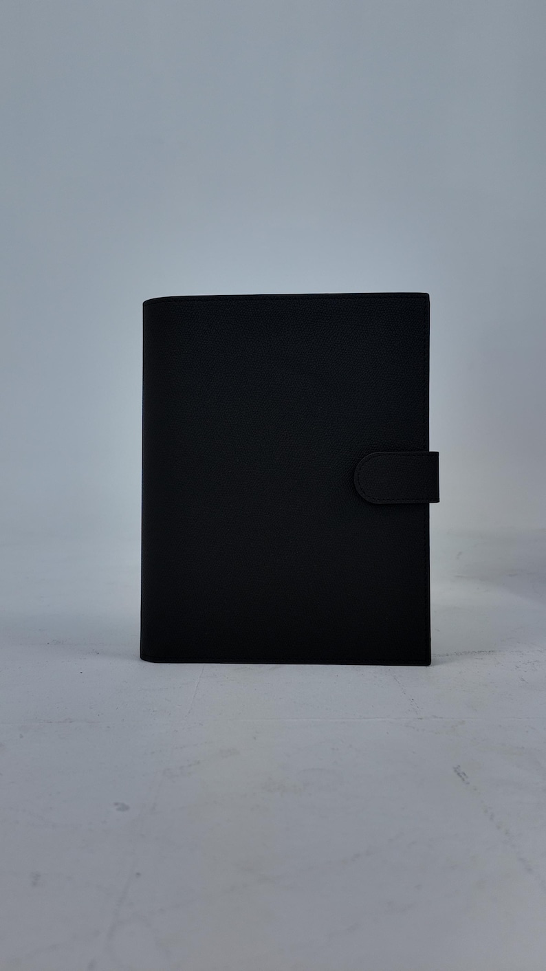 Peut inclure: Un journal noir rectangulaire avec une couverture textur&eacute;e et une fermeture magn&eacute;tique. Le journal est pos&eacute; verticalement sur un fond uni gris clair. La couverture a une finition lisse et mate.