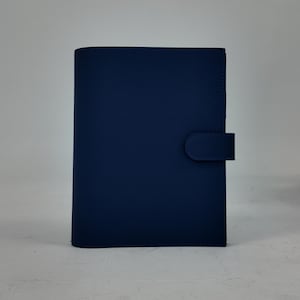 Peut inclure: Un journal en cuir bleu marine avec une surface textur&eacute;e. Le journal a une fermeture &agrave; rabat incurv&eacute;e sur le c&ocirc;t&eacute; droit. Le fond est gris clair uni.