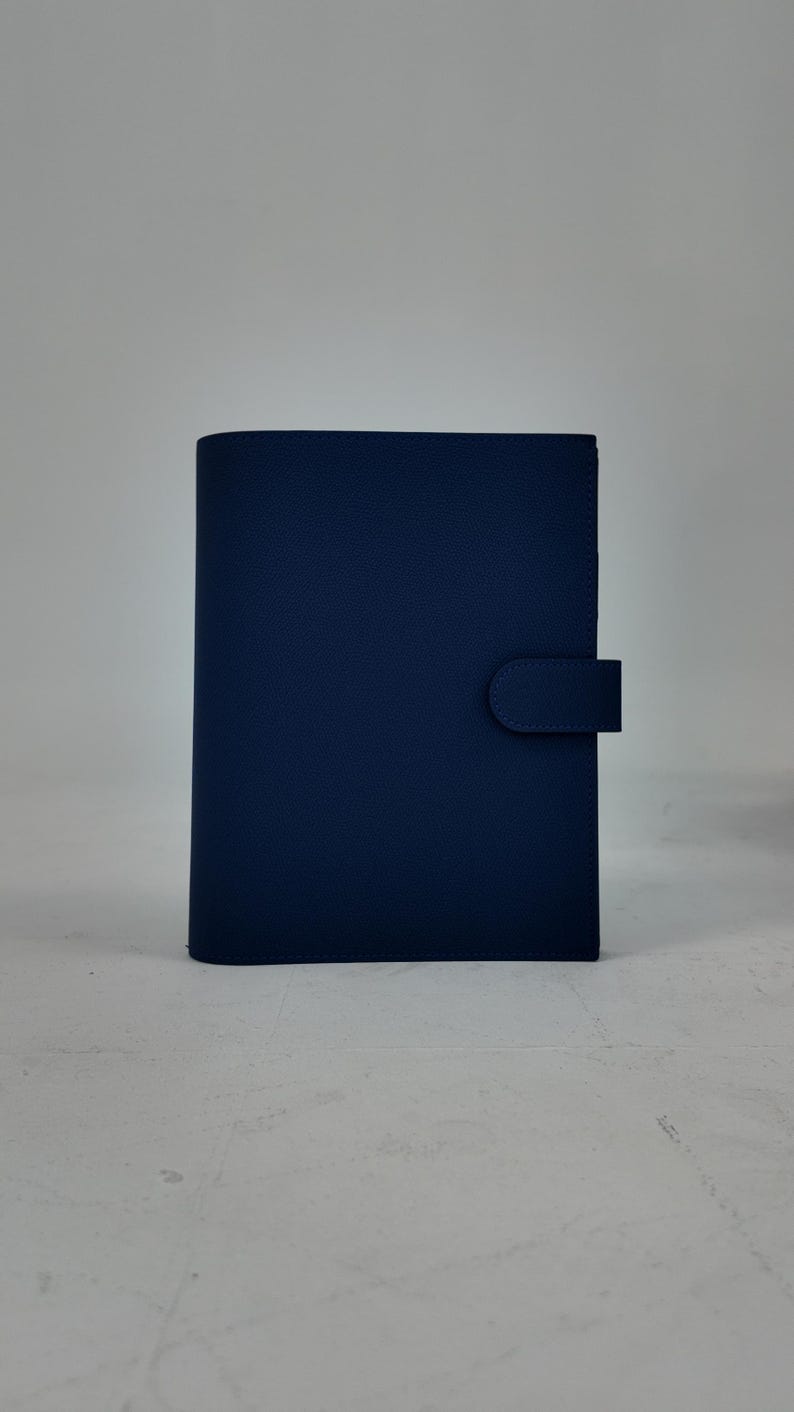 Peut inclure: Un journal en cuir bleu marine avec une surface textur&eacute;e. Le journal a une fermeture &agrave; rabat incurv&eacute;e sur le c&ocirc;t&eacute; droit. Le fond est gris clair uni.