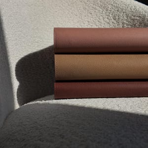 May include: Three stacked leather journals in shades of brown, from a light tan to a deep reddish-brown. The journals are neatly arranged, showcasing their textured surfaces and the subtle variations in color. The image is lit with soft, natural light.