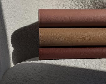 May include: Three stacked leather journals in shades of brown, from a light tan to a deep reddish-brown. The journals are neatly arranged, showcasing their textured surfaces and the subtle variations in color. The image is lit with soft, natural light.