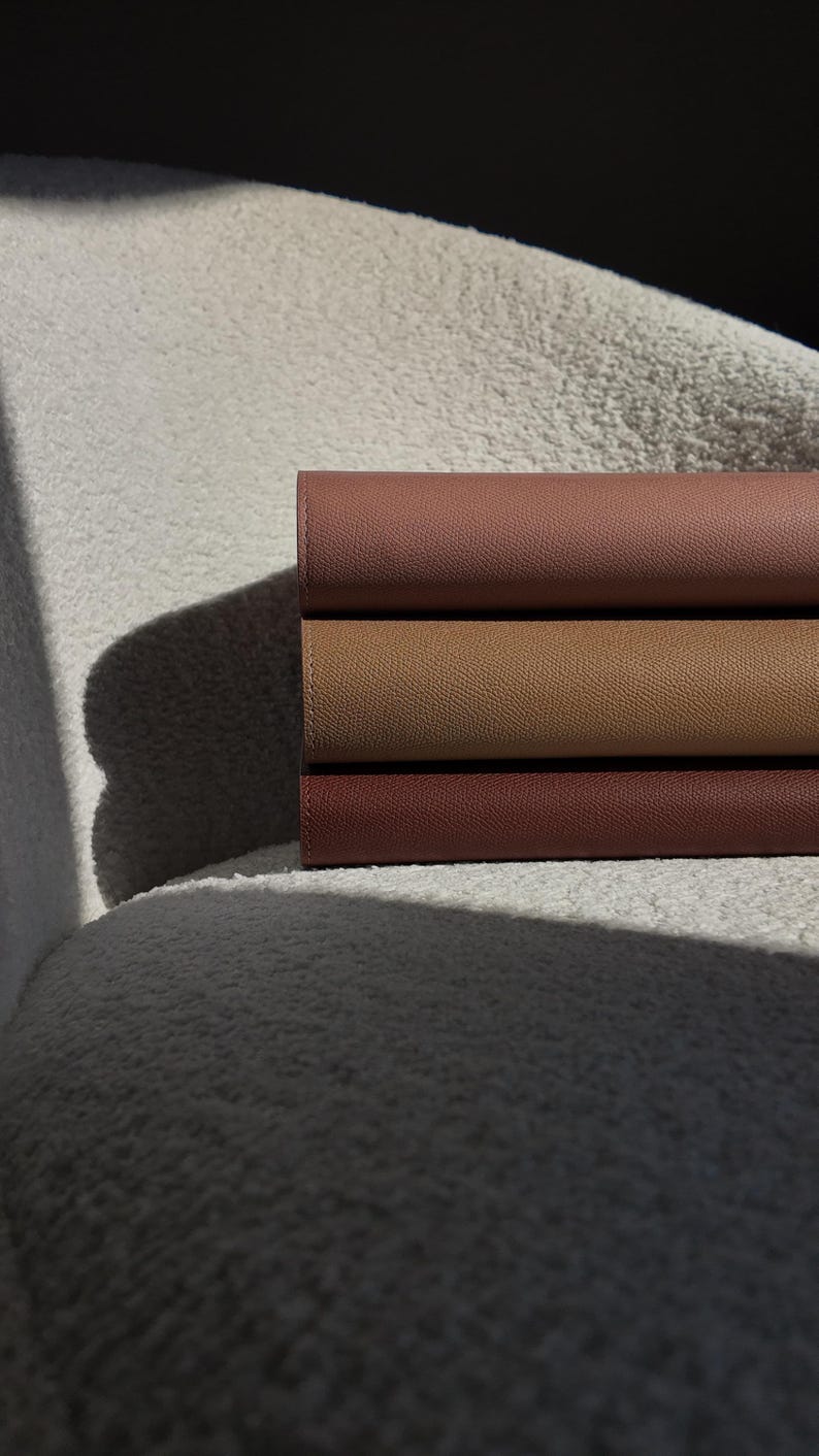 Peut inclure: Trois carnets en cuir empil&eacute;s dans des tons bruns, allant d'un beige clair &agrave; un brun rouge&acirc;tre fonc&eacute;. Les carnets sont soigneusement dispos&eacute;s, mettant en valeur leurs surfaces textur&eacute;es et les subtiles variations de couleur. L'image est &eacute;clair&eacute;e par une lumi&egrave;re naturelle douce.
