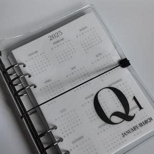 May include: A clear, plastic-covered planner with a black binding and elastic closure. The planner is open to a page displaying a 2025 calendar with the text "Q1 JANUARY-MARCH". The pages are white with grey text.