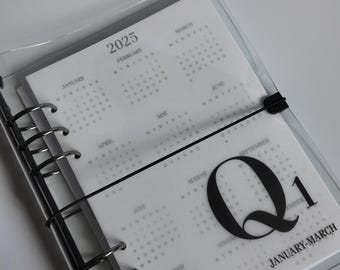 Może przedstawiać: Przezroczysty planer z czarną oprawą i elastycznym zapięciem. Planer jest otwarty na stronie z kalendarzem na rok 2025 z napisem "Q1 STYCZEŃ-MARZEC". Strony są białe z szarym tekstem.