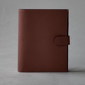 Peut inclure: Un journal en cuir marron avec une fermeture magn&eacute;tique. Le journal est rectangulaire et pr&eacute;sente une finition lisse et mate. La fermeture est une petite languette arrondie qui se fixe &agrave; l'avant. Le journal est ferm&eacute; et debout.
