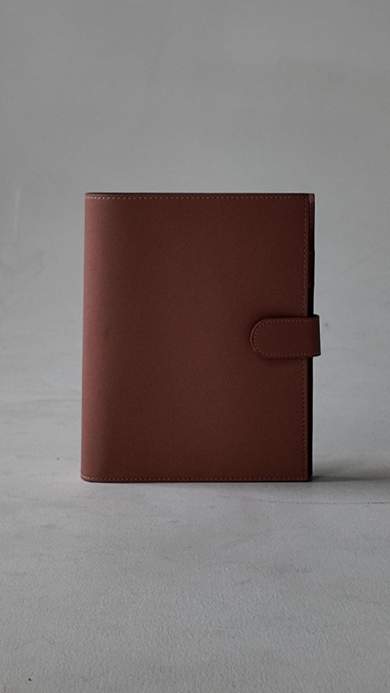 Peut inclure: Un journal en cuir marron avec une fermeture magn&eacute;tique. Le journal est rectangulaire et pr&eacute;sente une finition lisse et mate. La fermeture est une petite languette arrondie qui se fixe &agrave; l'avant. Le journal est ferm&eacute; et debout.