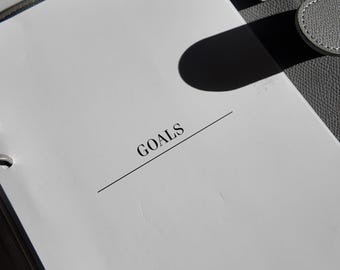 May include: A gray leather binder with a white page that reads "GOALS" in black text. The binder is open, revealing metal rings and a gray leather closure. The image is well-lit, with shadows adding depth.