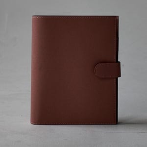 Peut inclure: Un journal en cuir marron avec une fermeture magnétique. Le journal est rectangulaire et présente une finition lisse et mate. La fermeture est une petite languette arrondie qui se fixe à l'avant. Le journal est fermé et debout.