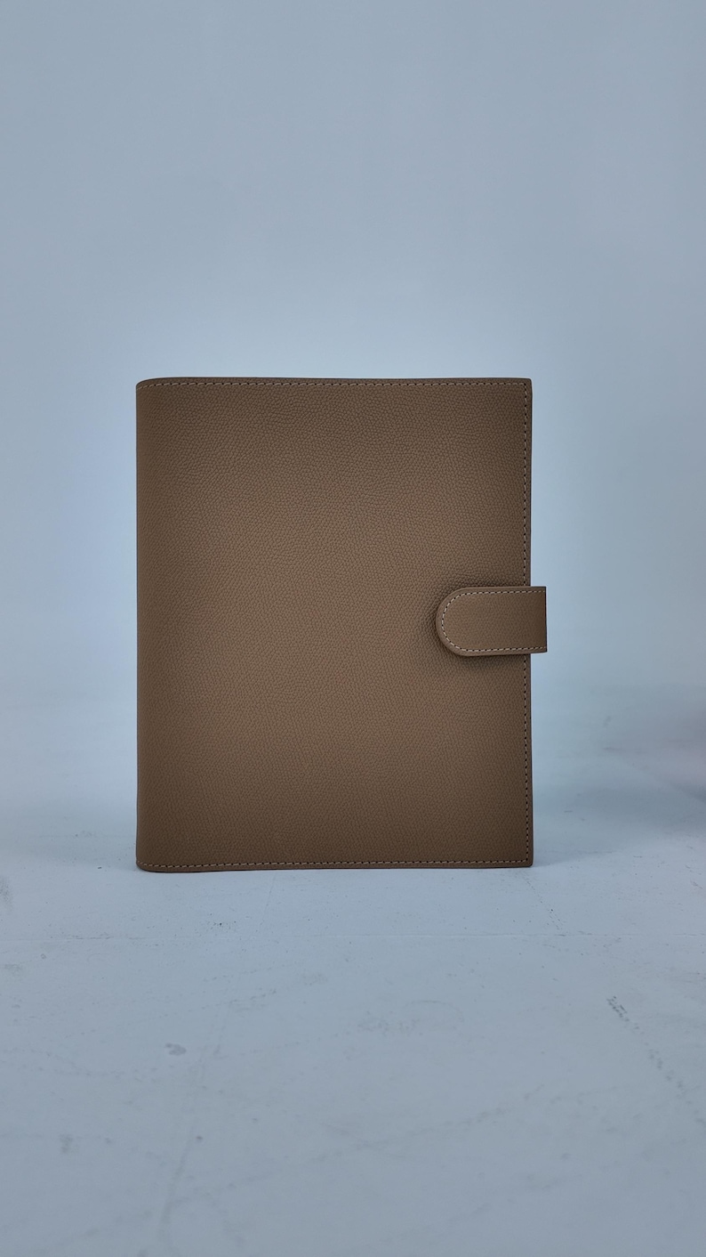 Peut inclure: Un agenda rectangulaire de couleur beige avec une fermeture &agrave; languette incurv&eacute;e. L'agenda est fait d'un mat&eacute;riau lisse et a une bordure cousue. La couverture est de couleur unie et neutre, adapt&eacute;e &agrave; divers usages.