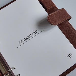 Peut inclure: Un classeur en cuir marron avec une page blanche sur laquelle est inscrit le mot "PRODUCTIVITÉ" en noir. Le classeur est doté d'un mécanisme à anneaux en métal argenté et d'une fermeture à lanière marron. Un petit logo est visible sur la page.