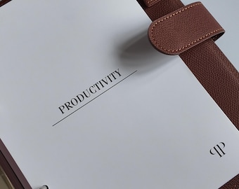 Könnte beinhalten: Ein brauner Lederordner mit einer weißen Seite, auf der in schwarzer Schrift "PRODUKTIVITÄT" steht. Der Ordner hat einen silbernen Metallringmechanismus und einen braunen Riemenverschluss. Ein kleines Logo ist auf der Seite sichtbar.