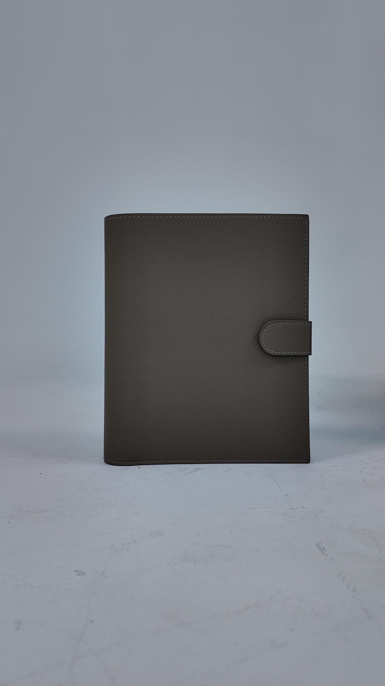 Peut inclure: Un journal en cuir gris avec une fermeture &agrave; bouton-pression. La couverture est de couleur grise unie et sourde avec une surface textur&eacute;e. Le journal est rectangulaire et semble ferm&eacute;, avec une fermeture &agrave; languette sur le c&ocirc;t&eacute; droit.