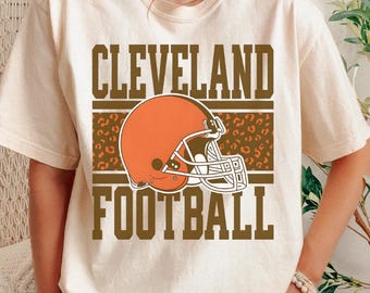 May include: Cream-colored t-shirt featuring the words "CLEVELAND FOOTBALL" in brown block letters. An orange football helmet is centered above a brown leopard print stripe. The shirt is short-sleeved.