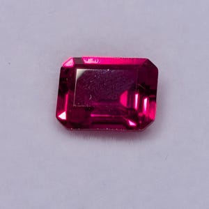 May include: A vibrant, rectangular-cut, magenta gemstone displayed in a white jewelry box. The gemstone has a faceted surface, reflecting light. The box is open, revealing the gem against a white, cushioned interior. This image showcases a precious stone.