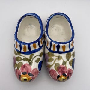 May include: Pair of ceramic clogs, hand-painted with floral and leaf designs in pink, green, and yellow on a white base. The clogs have blue and orange accents around the top rim.