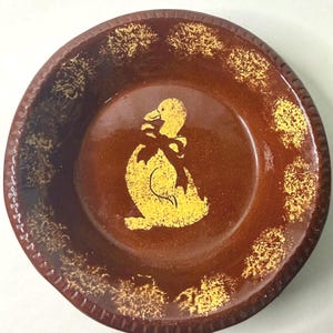 May include: A round brown ceramic bowl featuring a central yellow graphic of a duck wearing a bow. The rim of the bowl is decorated with a repeating yellow sponge-painted pattern, and the outer edge has a textured, ribbed design throughout.