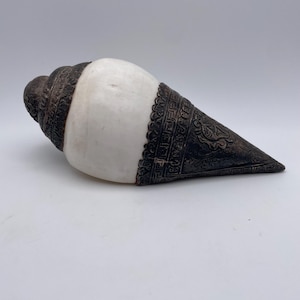 May include: A large, white conch shell with ornate, dark brown metalwork around the edges. The metalwork features intricate patterns and designs. The shell is a natural, organic shape, and the metal adds a decorative element.