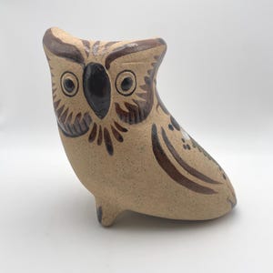 May include: A ceramic owl figurine in shades of tan and brown. The owl has large, round eyes, a dark beak, and detailed feather markings. The figurine is standing upright with a slightly tilted head, showcasing a handcrafted aesthetic.